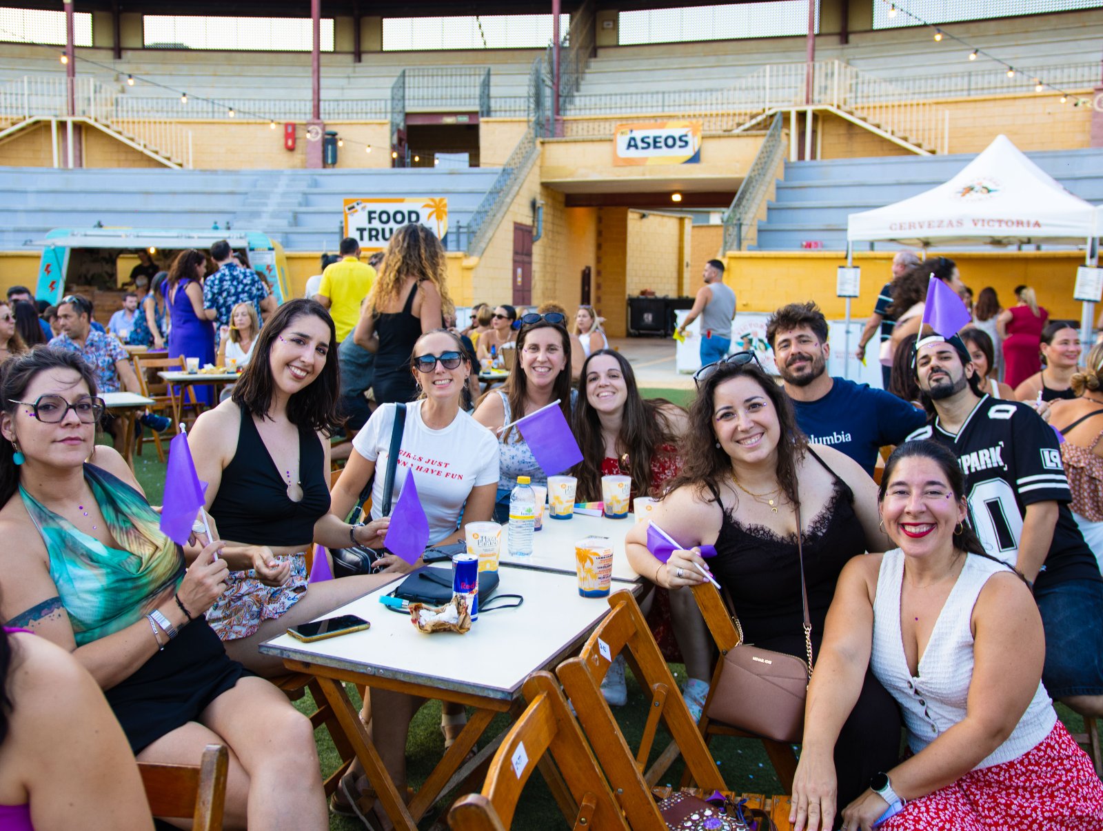Grupo de amigas sentadas en una mesa del ruedo con banderines morados y copas, junto a los food trucks de Plaza Paraíso en Furor The Show
