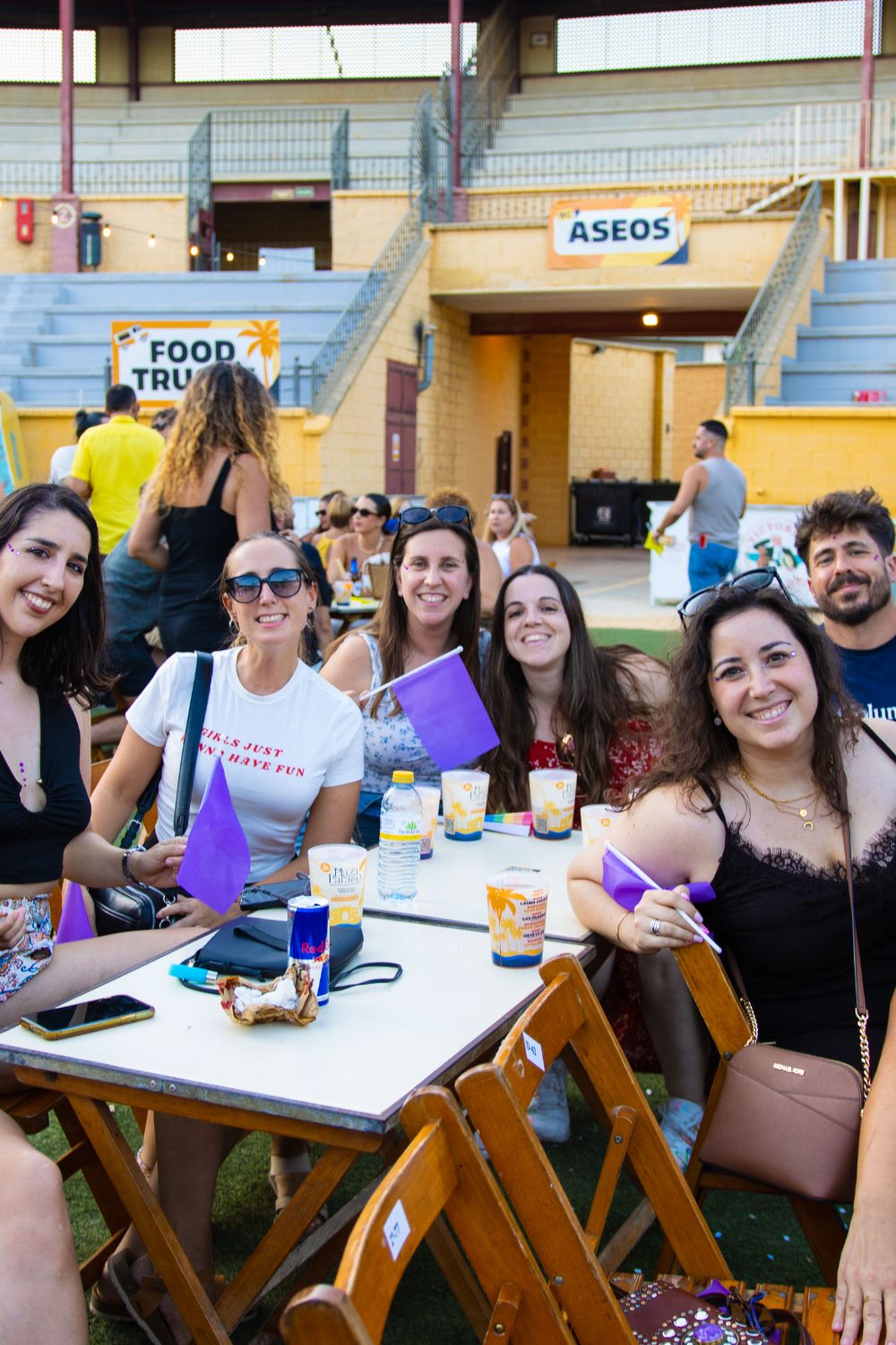 Grupo de amigas sentadas en una mesa del ruedo con banderines morados y copas, junto a los food trucks de Plaza Paraíso en Furor The Show