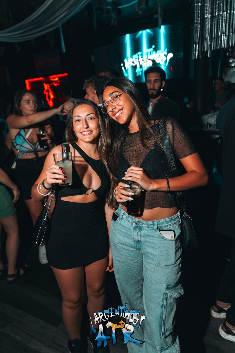 Dos amigas posando con sus consumiciones delante del cartel luminoso de ¡Argentinos ATR! en la fiesta
