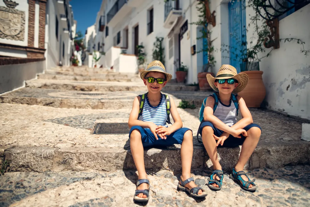 Dos niños con sombreros de paja y gafas de sol sentados en las escaleras de un pueblo blanco andaluz en Málaga, plan de verano perfecto para familias en la Costa del Sol